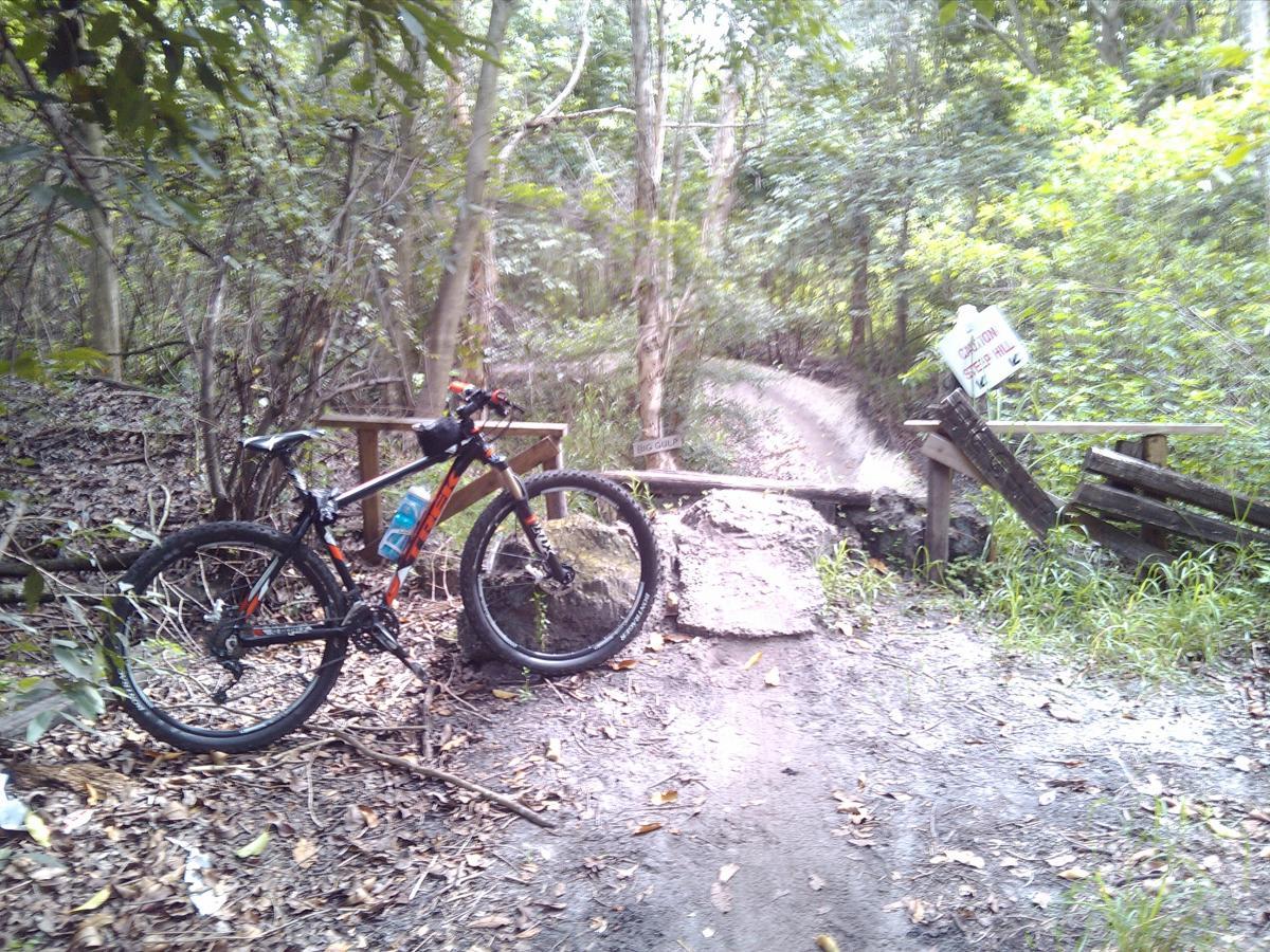 A mountain bike resting on a gravel path in a dense forested area. In the background, a sign indicates caution and alerts to a nearby hill. The scene is surrounded by lush greenery and scattered leaves on the ground. Markham Park mountain bike trail.