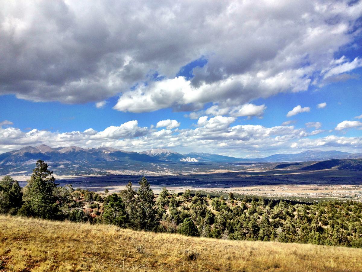 A panoramic view of a mountainous landscape under a partly cloudy sky, featuring lush green trees in the foreground and distant peaks in the background. The scene captures the natural beauty of rolling hills and valleys, with varied cloud formations adding depth to the sky. Rainbow Trail: Methodist Mountain Thd to Bear Creek Thd mountain bike trail.