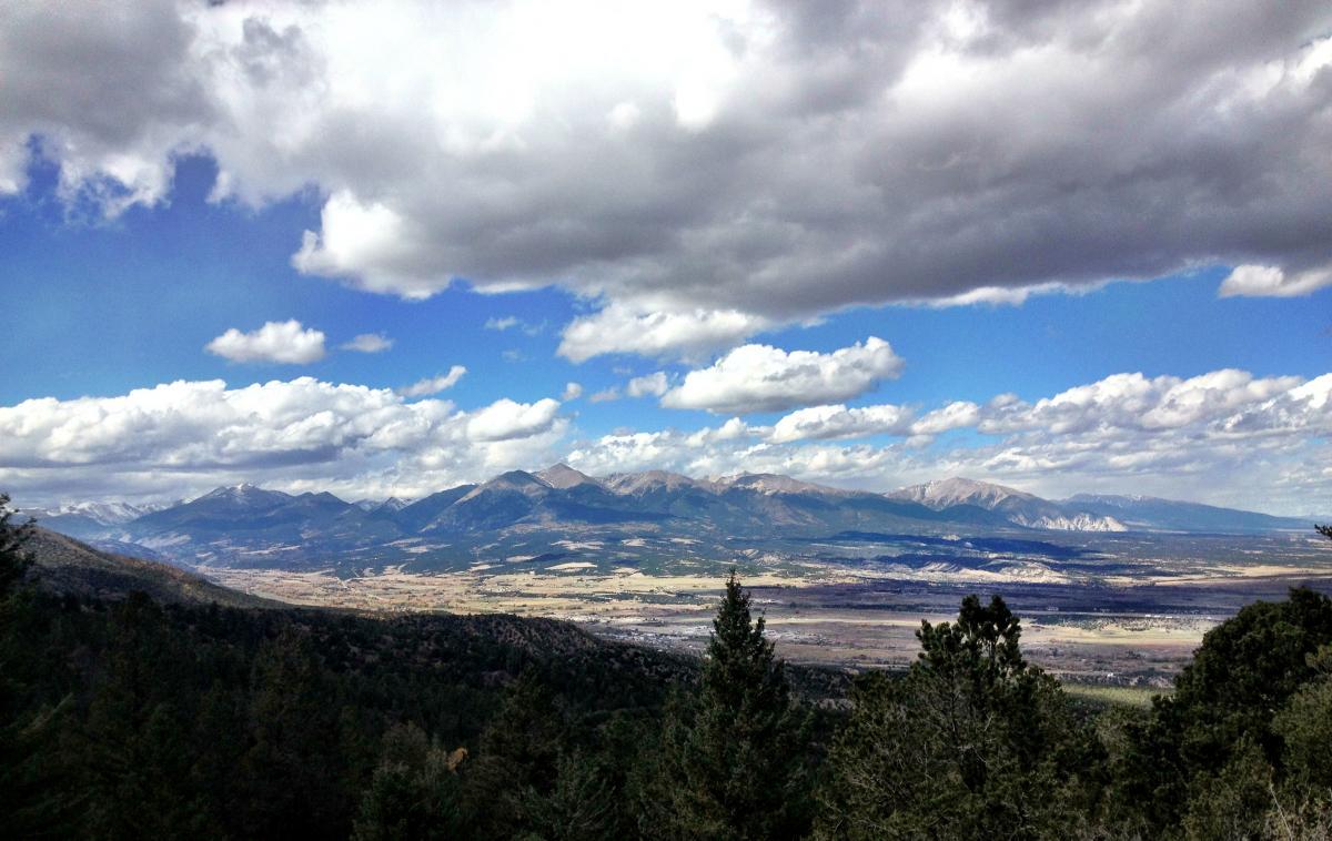 A panoramic view of a mountainous landscape under a partly cloudy sky, featuring rolling hills, rugged peaks, and a vast valley filled with greenery. The foreground includes dense trees, while the background showcases mountains with varying elevations and a mix of sunlight and shadows across the terrain. Rainbow Trail: Methodist Mountain Thd to Bear Creek Thd mountain bike trail.