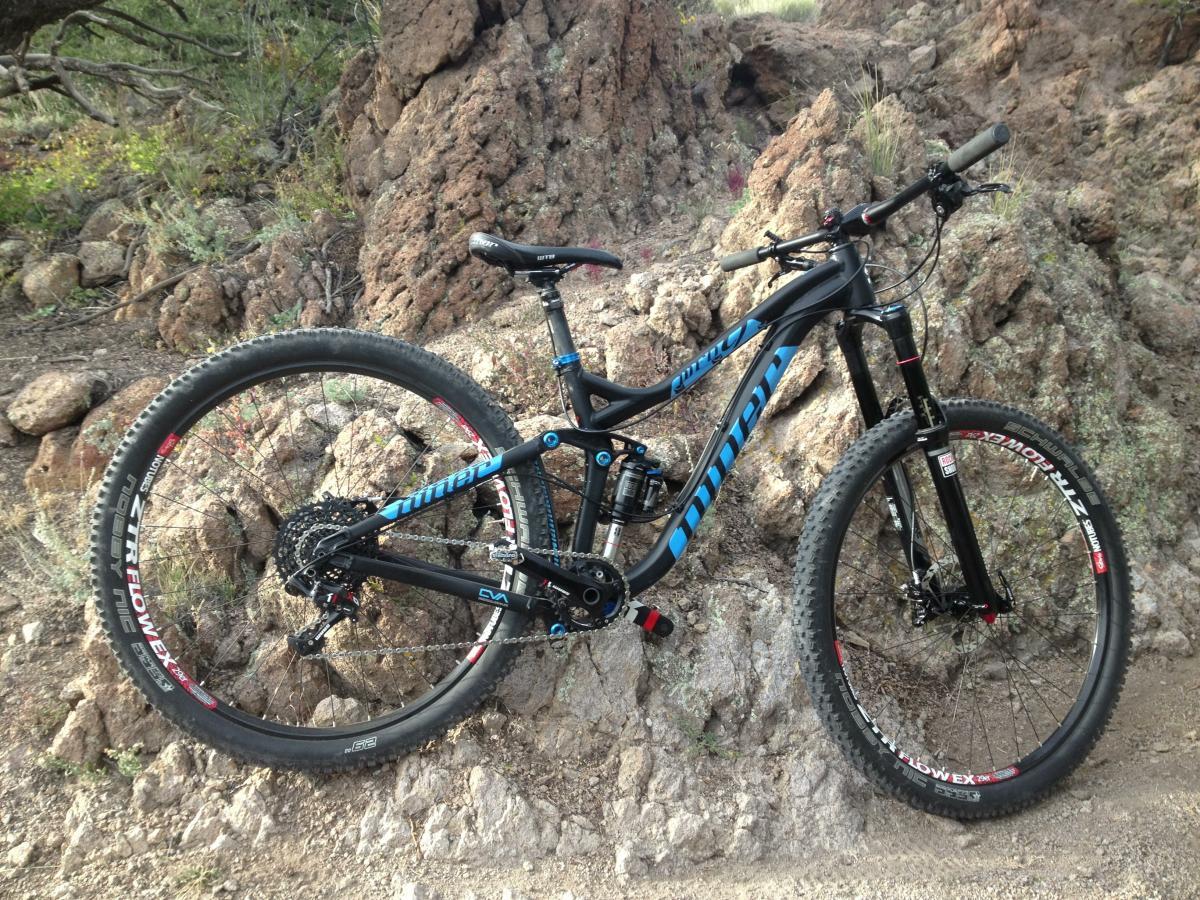 Niner W.F.O. 9: A black and blue mountain bike leaning against a rocky terrain, surrounded by greenery and dirt. The bike features thick tires and a strong frame, ideal for off-road riding.