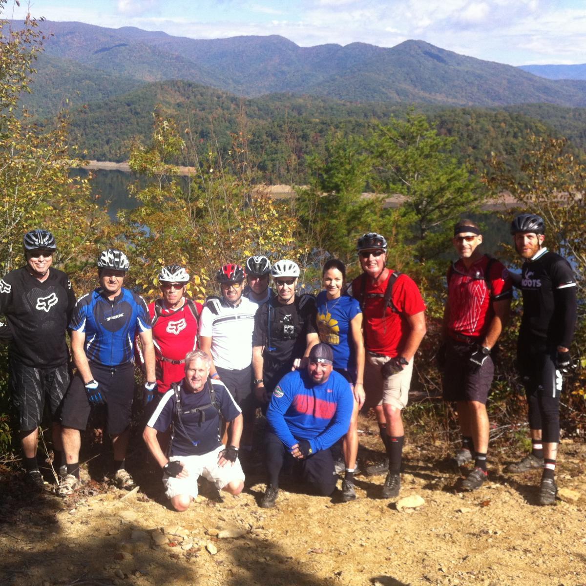A group of eleven mountain bikers poses for a photo outdoors, surrounded by trees and distant mountains. They are wearing various cycling gear, including helmets and jerseys in a mix of colors. The landscape features a body of water in the background, and the sky is partly cloudy with blue patches. The scene suggests a day of cycling and enjoying nature.