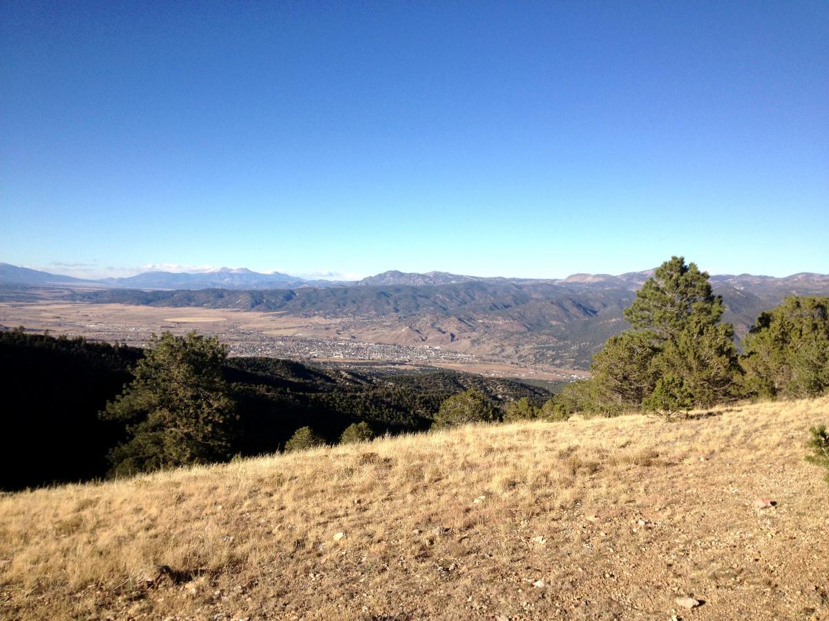 A scenic overlook of a mountainous landscape under a clear blue sky, featuring rolling hills and valleys. Green trees are visible in the foreground, with distant mountains and a valley stretching out below. The scene conveys a sense of tranquility and natural beauty. Rainbow Trail: Methodist Mountain Thd to Bear Creek Thd mountain bike trail.