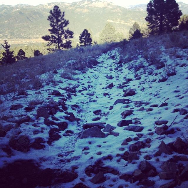 A winding dirt path covered in patches of snow and surrounded by rocky terrain, leading through a hilly landscape with distant mountains and sparse trees in the background. Rainbow Trail: Hwy 285 to Methodist Mountain Thd mountain bike trail.