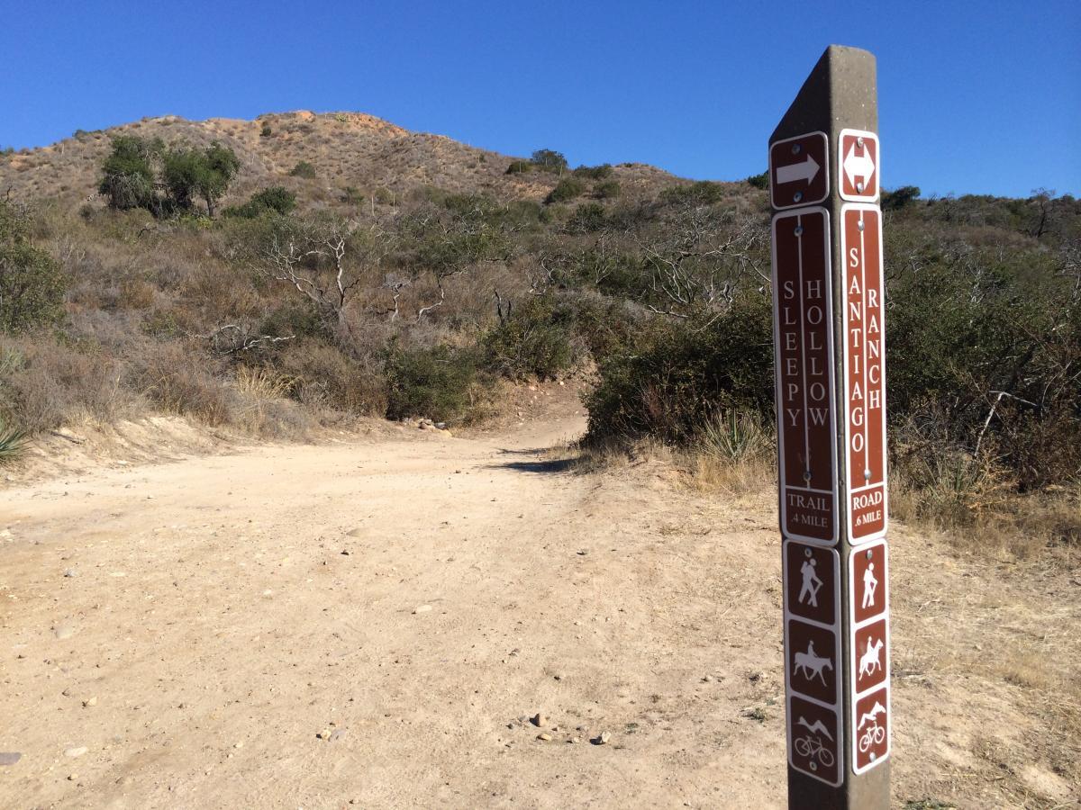 A trail sign indicating directions for the Sleepy Hollow trail and Santiago Road, located in a dry, hilly area with sparse vegetation under a clear blue sky. The sign features symbols for hiking, horseback riding, and biking, with distances marked for each destination. Santiago Ranch Rd mountain bike trail.
