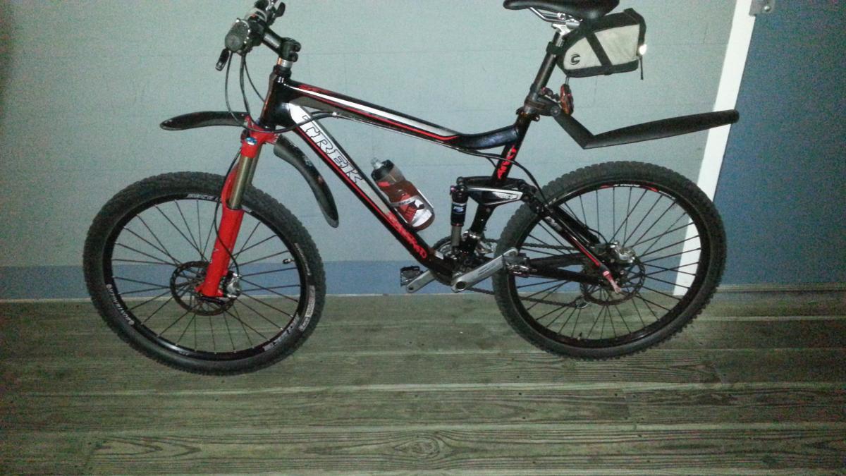 Trek Fuel EX 8: A black and red mountain bike parked on a wooden floor against a light-colored wall. The bike features thick tires, a water bottle holder, and a small storage pouch attached to the handlebars.