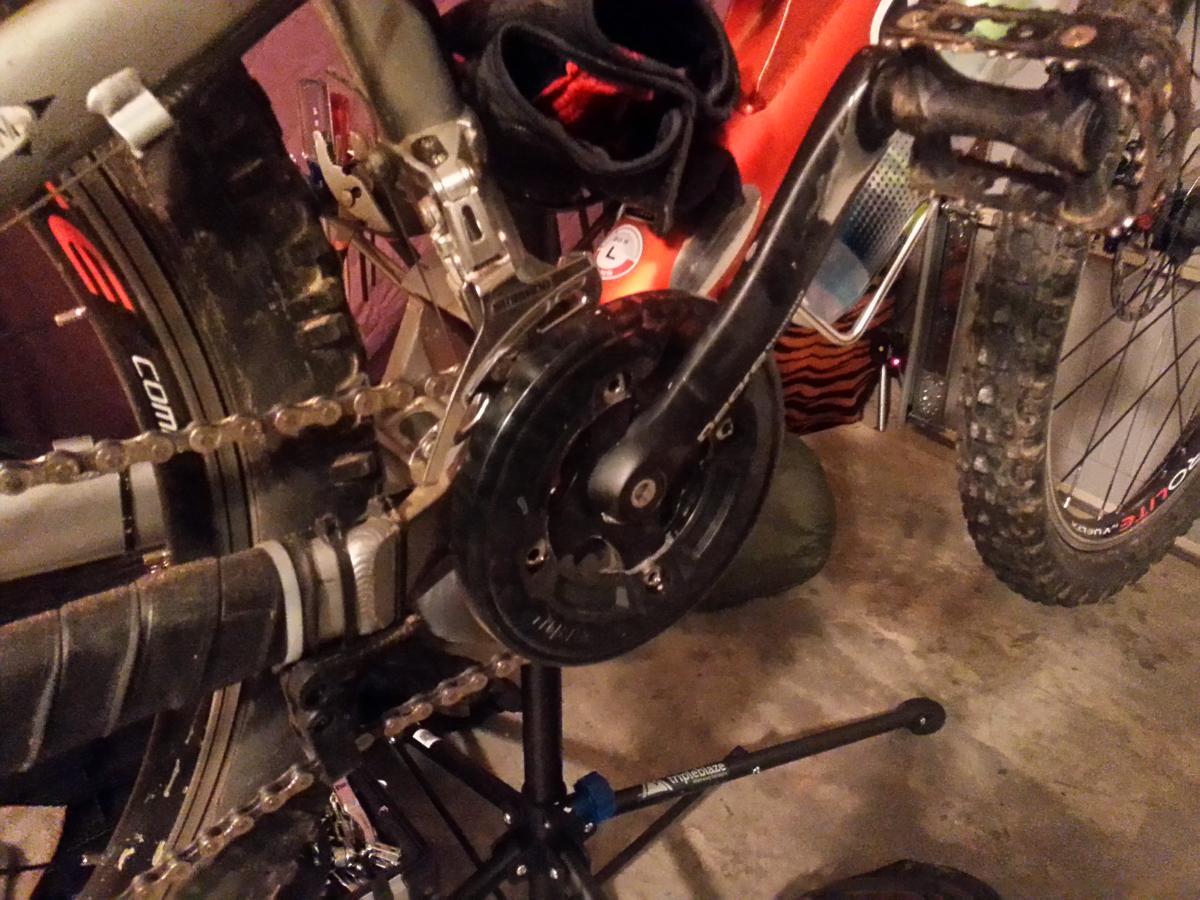 Shimano Saint Bashguard: A close-up image of a bicycle's drivetrain, focusing on the crankset, chain, and rear derailleur. The bicycle is positioned on a repair stand in a garage. The crank arm is black, with a chain running through a large black chainring. The rear wheel, partially visible, has a knobby tire and appears to have dirt on it. Tools and various items are noticeable in the background.