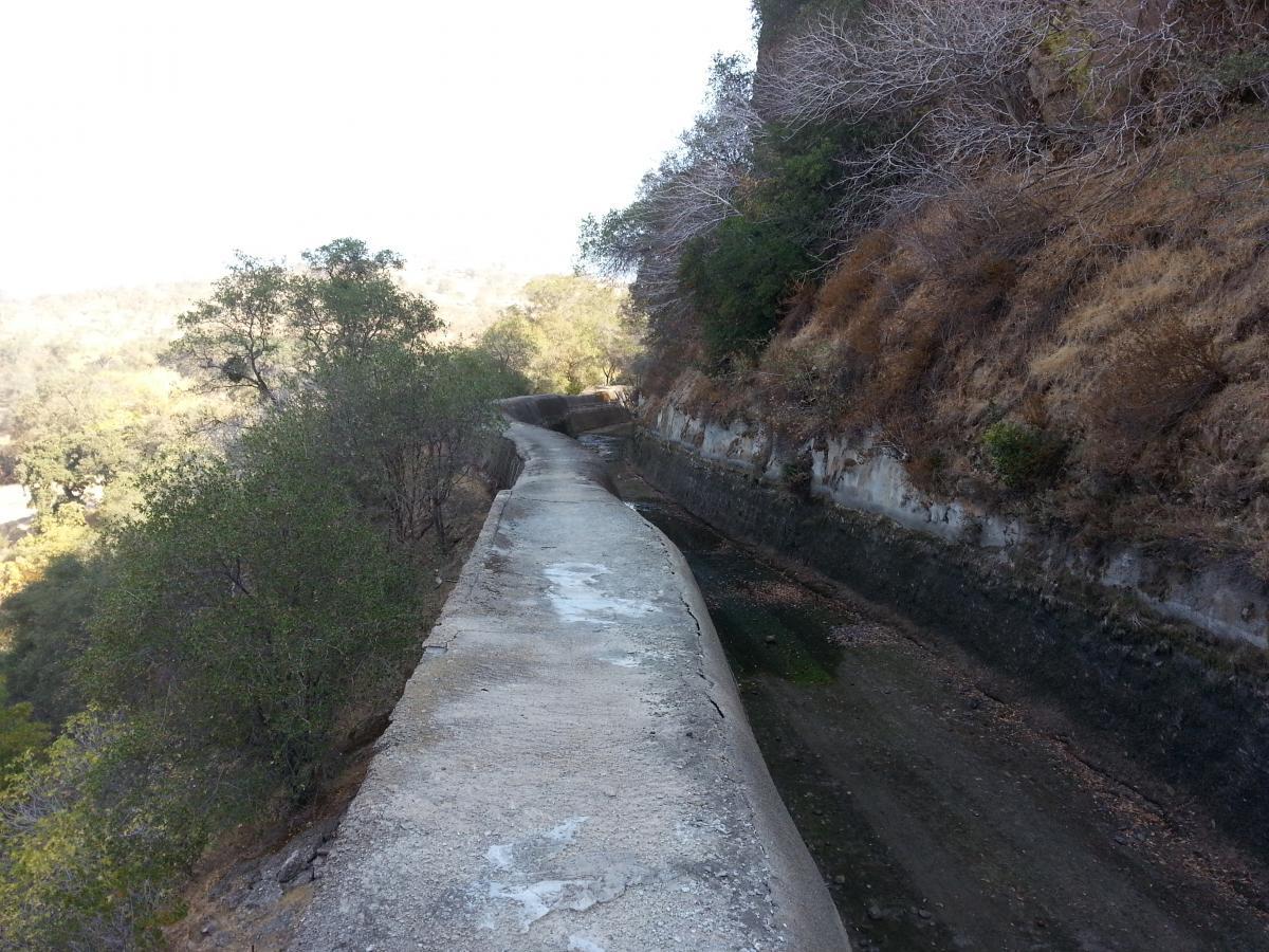A narrow, weathered concrete pathway lined with vegetation runs alongside a shallow, stagnant waterway. The path leads into the distance, bordered by rocky hills and sparse trees, under a bright sky. Knights Ferry Canal mountain bike trail.