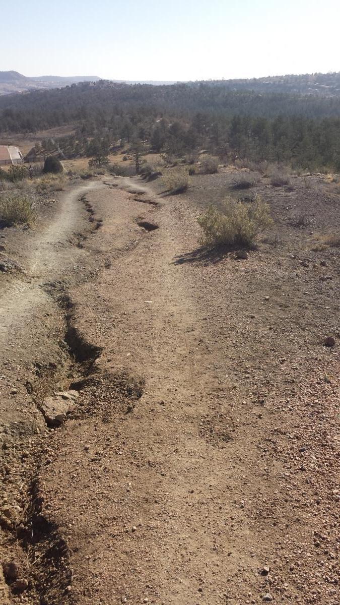 A winding dirt path leads through a rugged landscape, flanked by sparse vegetation and small bushes. The terrain is rocky and dry, with light-colored soil and a few visible tire tracks. In the background, rolling hills and a clear blue sky can be seen, indicating a vast, open area. Ute Valley Park mountain bike trail.
