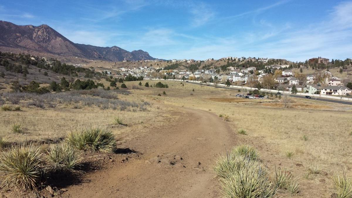 A dirt trail winds through a dry, grassy landscape, leading towards a residential area nestled at the base of mountains under a clear blue sky. Sparse vegetation and a few low shrubs dot the scene, while houses are visible in the background along a road. Ute Valley Park mountain bike trail.
