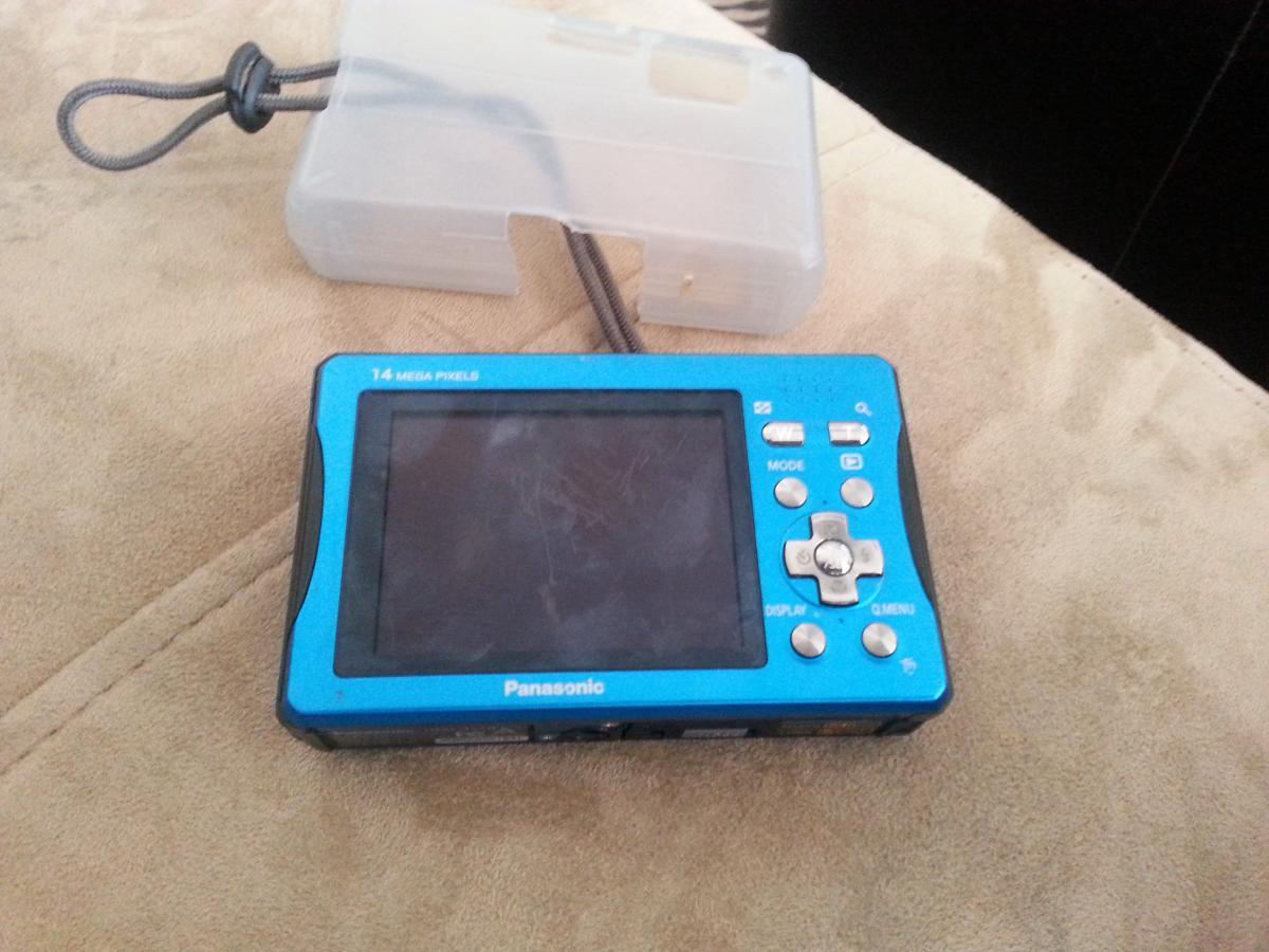 Panasonic Lumix DMC TS10: A blue Panasonic digital camera featuring a 14-megapixel display. The camera has various buttons for settings and functions, and is positioned on a soft surface with a light-colored background. A clear protective case is partially visible alongside the camera.