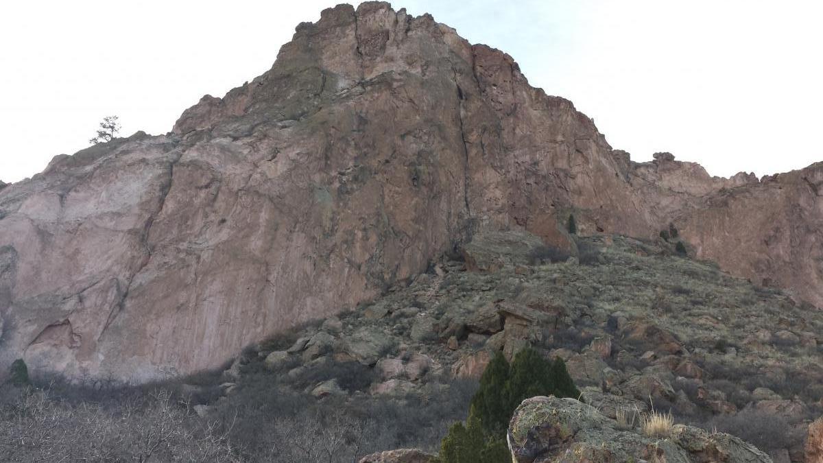 A steep rock formation rises majestically from a hillside, surrounded by sparse vegetation and patches of grass. The rocky landscape features rugged textures and varying colors of stone, with trees visible at the summit and on the slopes. The sky above is overcast, adding a subdued ambiance to the scene. Garden of the Gods: Ute Trail mountain bike trail.