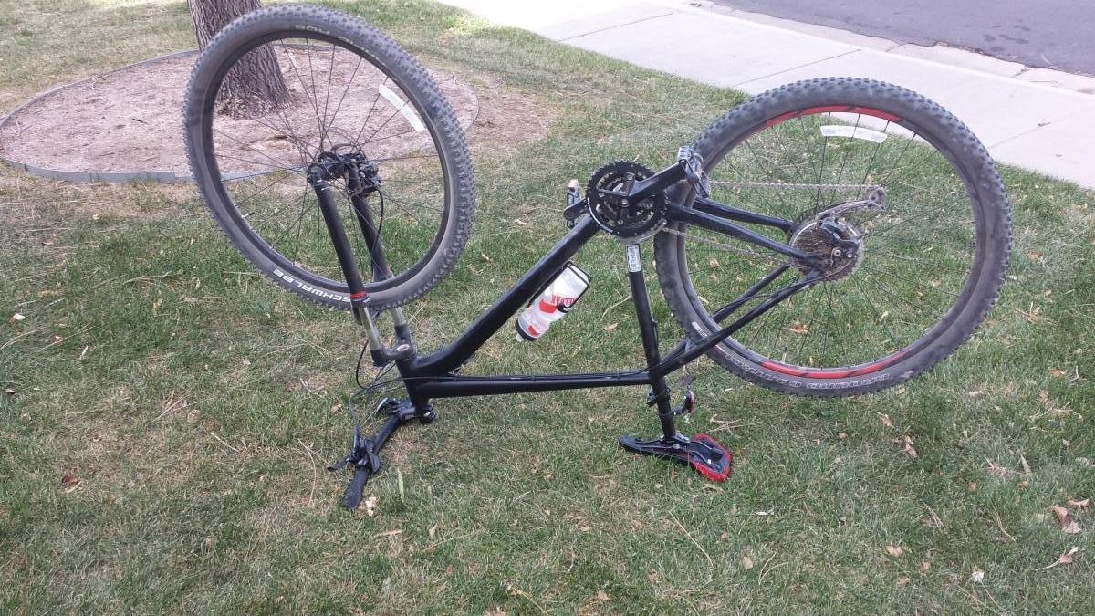 An upside-down black mountain bike resting on the grass, revealing its wheels and frame, with a water bottle mounted on the frame. A sidewalk and tree are visible in the background.