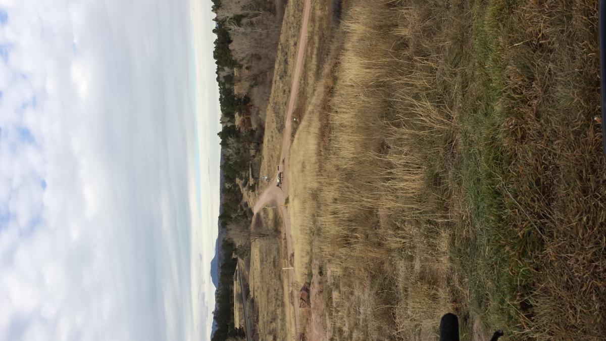 A scenic view of rolling hills and dirt paths, with tall grasses in the foreground and a cloudy sky overhead. The landscape includes sparse trees and winding trails, suggesting a tranquil outdoor setting. Santa Fe Trail mountain bike trail.