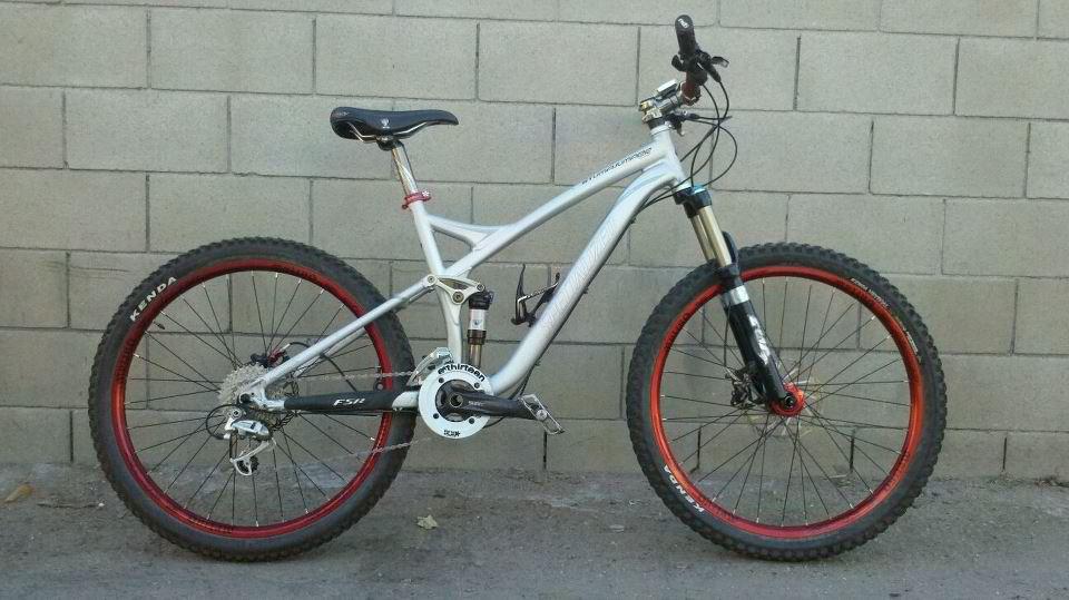 Specialized Stumpjumper: A silver mountain bike with red accents, featuring wide tires and a dual suspension frame, leaning against a grey concrete wall. The bike has disc brakes and a standard gear setup, showcasing its rugged design suitable for off-road riding.
