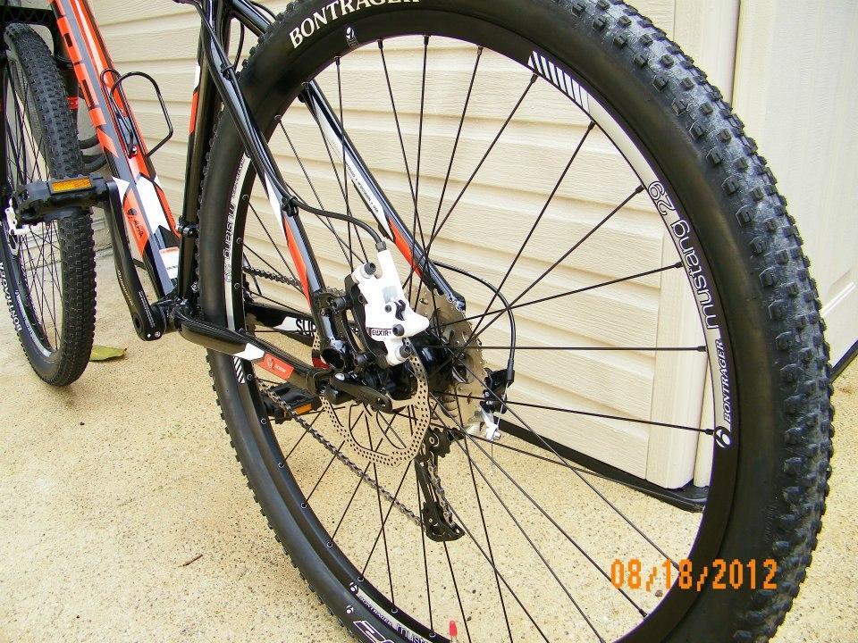 Trek Superfly AL: Close-up view of the rear wheel and drivetrain of a mountain bike, featuring a Bontrager tire, disc brake, and visible gears. The bike is positioned against a light-colored wall, with the focus on the components and mechanics of the bike.