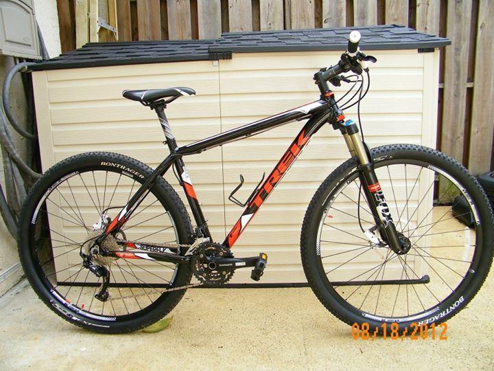 Trek Superfly AL: A black and red Trek mountain bike leaning against a storage shed, with visible features including thick tires, a front suspension fork, and various gears. The bike is positioned on a concrete surface, with a wooden fence in the background. The date is noted at the bottom of the image.