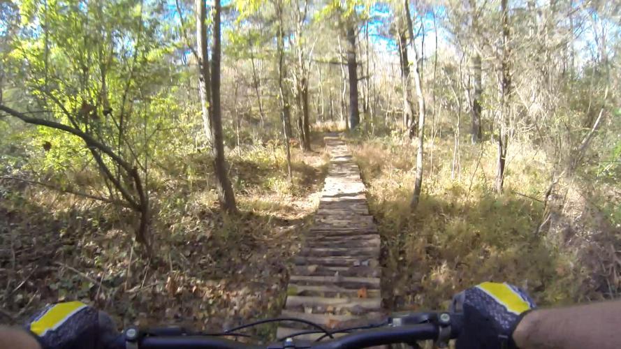 A view from the handlebars of a mountain bike navigating a wooden plank trail through a sunlit forest. The trail is surrounded by trees and undergrowth, with a clear blue sky visible above. Nockamixon State Park mountain bike trail.