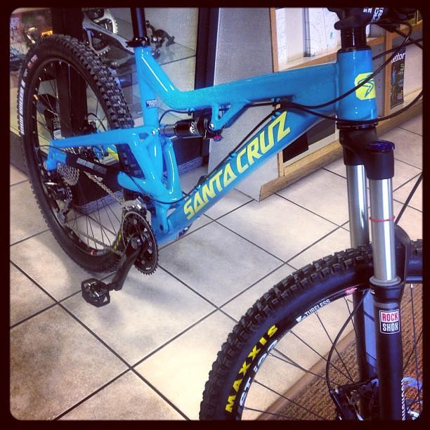 Santa Cruz Heckler: A close-up view of a blue Santa Cruz mountain bike displayed in a shop. The bike features a sleek frame, prominent branding, and a high-quality suspension system. The front wheel shows a Maxxis tire and a RockShox fork, while the background includes shelves and items typical of a bike shop.