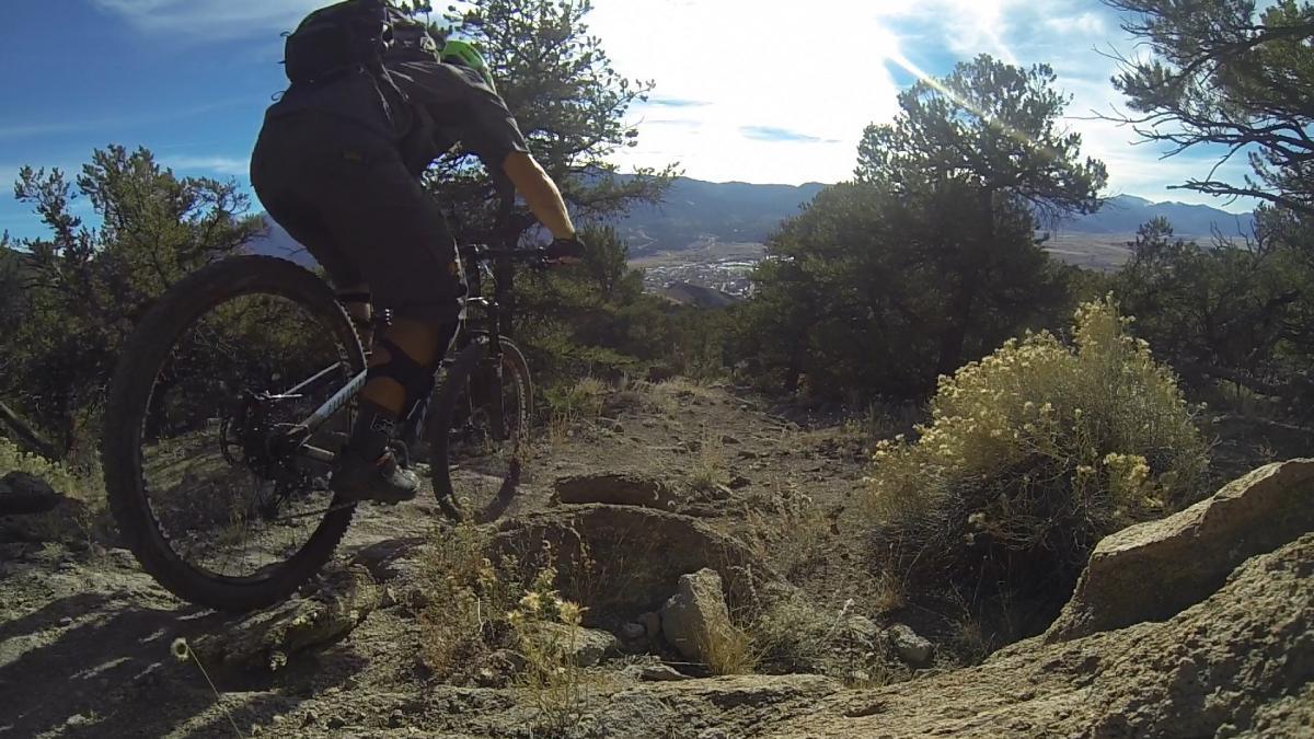 A mountain biker is mid-jump while riding down a rocky trail surrounded by trees and shrubs, with a scenic view of the valley and mountains in the background under a clear sky. Unkle Nazty mountain bike trail.