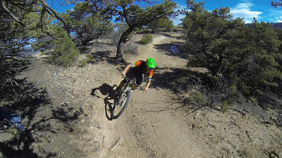 A mountain biker wearing a bright green helmet and a colorful jersey is leaning into a turn on a dirt trail surrounded by sparse vegetation and trees. The scene captures the rider in motion against a clear blue sky, highlighting the rugged outdoor terrain. Little Rainbow mountain bike trail.