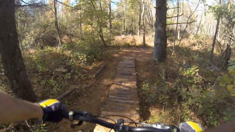 A view from the handlebars of a mountain bike navigating a wooden plank bridge along a forest trail, surrounded by trees and autumn foliage. Sunlight filters through the leaves, illuminating the path ahead. Nockamixon State Park mountain bike trail.