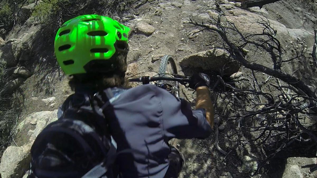A mountain biker navigating a rocky trail, wearing a bright green helmet and a black shirt, with a focus on the terrain ahead. The scene includes rough, natural ground with scattered rocks and sparse vegetation. Unkle Nazty mountain bike trail.