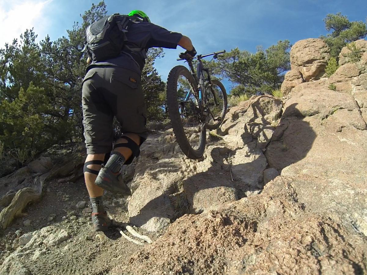 A person in athletic wear, including knee pads, is pushing a mountain bike up a rocky incline surrounded by trees and blue sky. The terrain consists of uneven, rocky surfaces, highlighting the challenges of off-road biking. Unkle Nazty mountain bike trail.