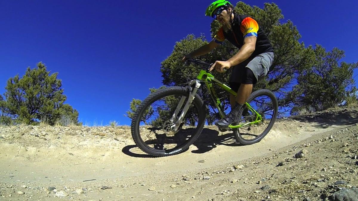 A mountain biker wearing a colorful jersey and helmet rides along a dirt trail, surrounded by sparse vegetation and a clear blue sky. The biker is leaning into a turn on a rocky path, showcasing an active outdoor scene. Little Rainbow mountain bike trail.
