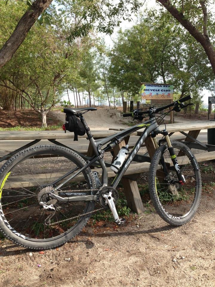 Scott Spark 950: A mountain bike parked next to a wooden picnic table in a wooded area, with a sign for the Virginia Key Bicycle Club visible in the background. The bike features a black frame, suspension components, and a water bottle attached to its frame. Trees and a sandy path are visible in the surrounding area.