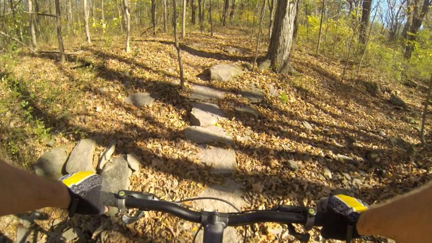 A view from the handlebars of a mountain bike riding along a rocky trail covered with fallen leaves, surrounded by trees in a forested area. Sunlight filters through the branches, casting shadows on the path. Nockamixon State Park mountain bike trail.