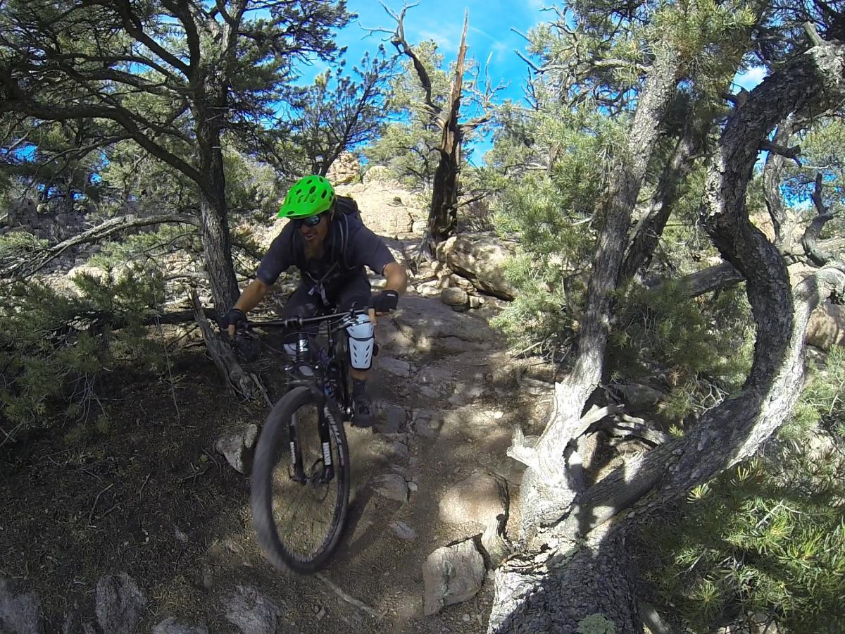 A mountain biker wearing a green helmet and protective gear navigates a rocky trail surrounded by trees under a clear blue sky. Unkle Nazty mountain bike trail.