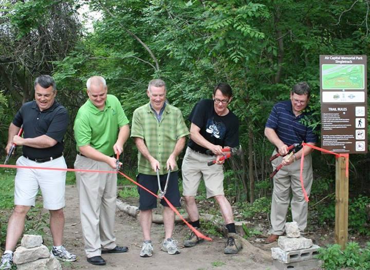 Five men stand in a line, each holding a pair of large scissors as they prepare to cut a red ribbon stretched across a trail entrance. The background features lush greenery and a sign detailing park rules and information about Air Capital Memorial Park. The men are wearing casual clothing in various colors and appear to be celebrating an event, possibly a trail opening. Air Capital Memorial Park mountain bike trail.