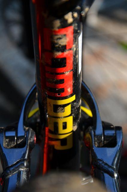 Specialized Camber 29: Close-up view of a bicycle frame with visible dirt and wear, featuring bold red and yellow branding elements. The image captures the intricate details of the bike's components and frame design.