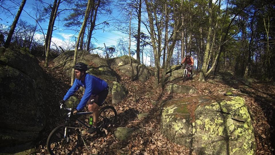 Two mountain bikers navigating a rocky trail in a wooded area, surrounded by trees and fallen leaves. One rider is in the foreground, wearing a blue shirt and a helmet, while the other is further back on the trail. The sky is blue, indicating a clear day. Raccoon Mountain Trail Network mountain bike trail.