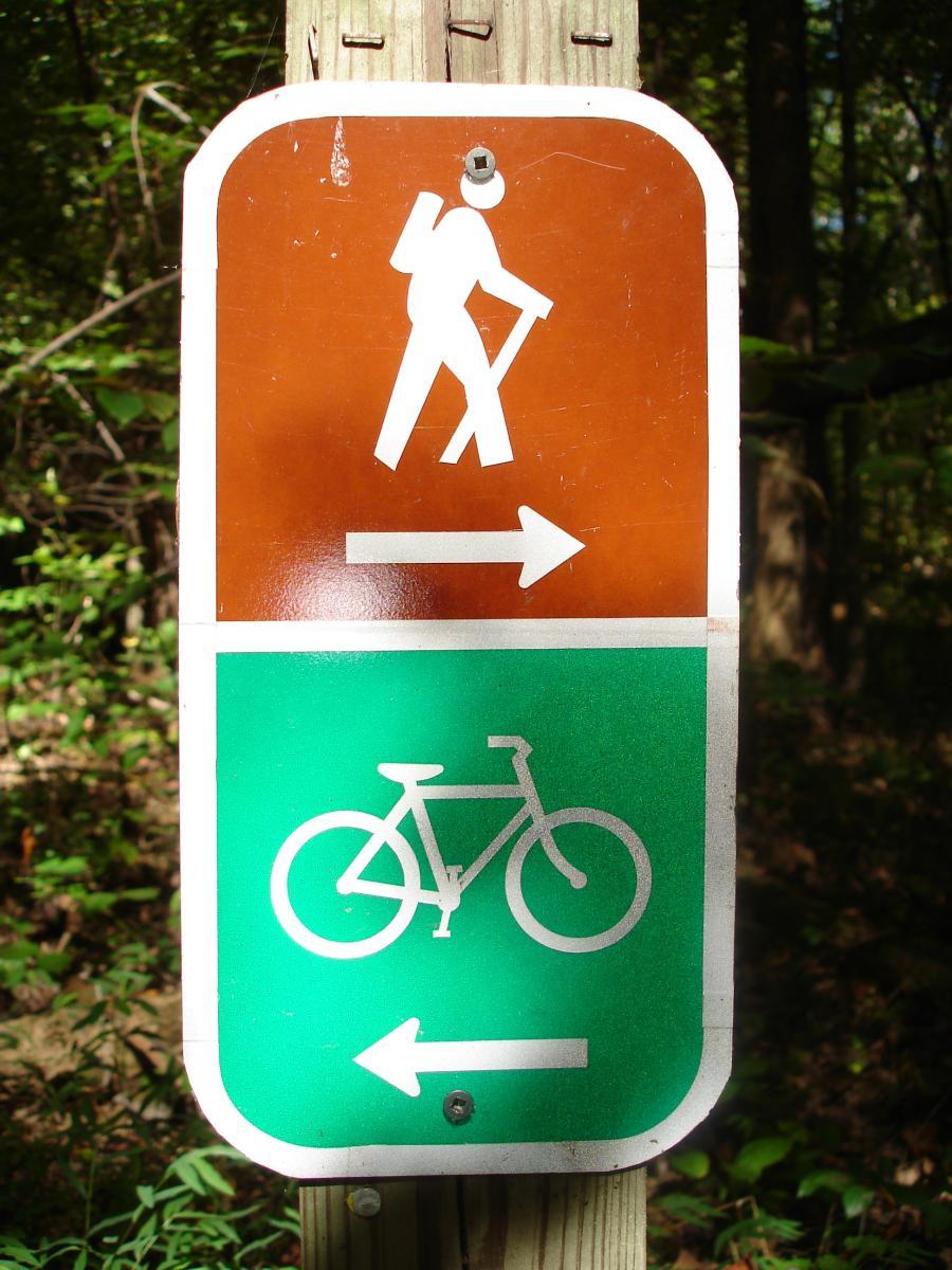 Sign indicating recreational trail directions: a hiker symbol pointing right on a brown background and a bicycle symbol pointing left on a green background. Itusi @ Lake Norman State Park mountain bike trail.
