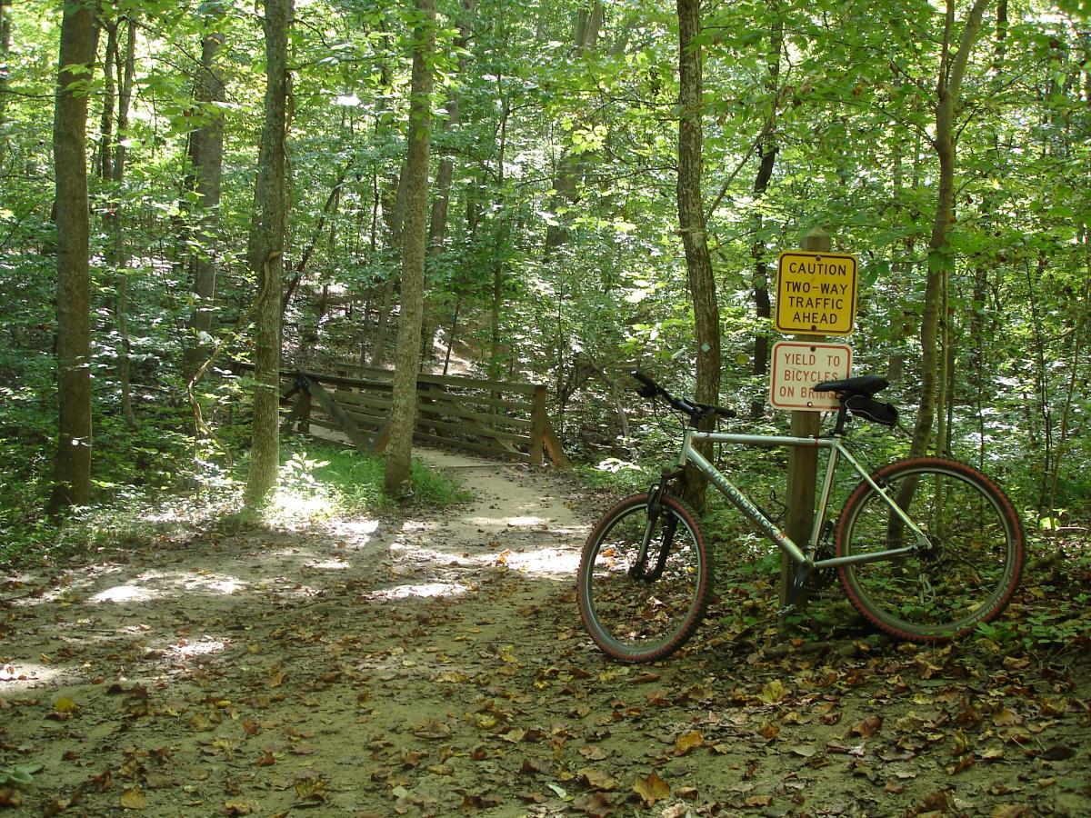 A mountain bike leaning against a sign in a wooded area, with trees lining a dirt path and a wooden bridge visible in the background. Signs indicate caution for two-way traffic and yield to bicycles on the bridge. Sunlight filters through the leaves, creating a dappled effect on the ground covered in fallen leaves. Itusi @ Lake Norman State Park mountain bike trail.