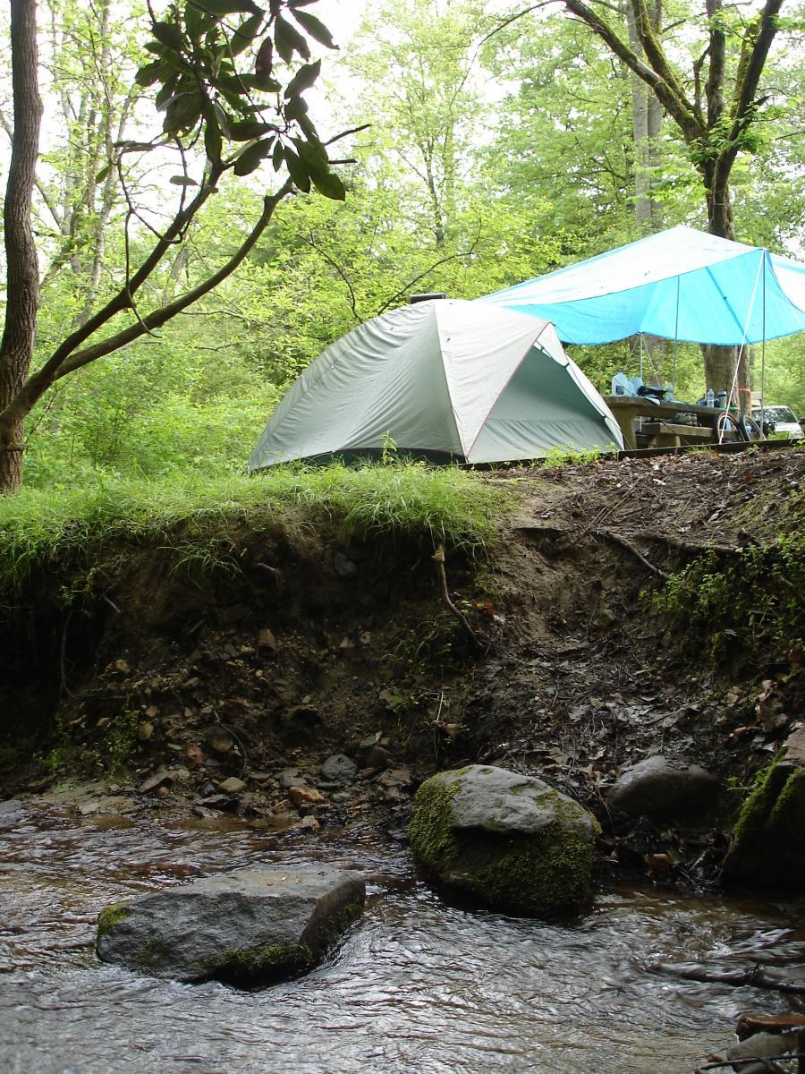 A camping scene showing a green tent and a blue canopy set up near a shallow stream, surrounded by lush green trees and foliage. The ground is uneven and features several moss-covered rocks in the water. Tsali Recreation Area mountain bike trail.
