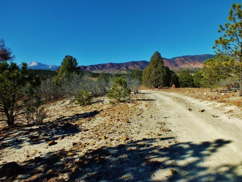 A dirt path winding through a sparse landscape, with small evergreen trees and rocky terrain. In the distance, majestic mountains are visible under a clear blue sky. Ute Valley Park mountain bike trail.