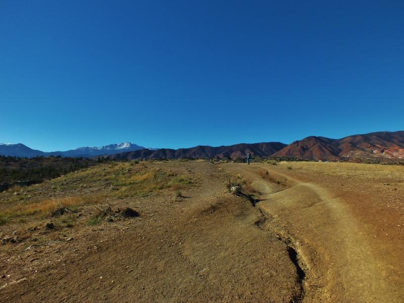 A scenic landscape featuring a winding dirt path leading through a hilly terrain, with a person walking in the distance. The background showcases majestic mountains, some capped with snow, under a clear blue sky. The foreground includes patches of dry grass and rocky soil, suggesting a natural outdoor setting. Ute Valley Park mountain bike trail.