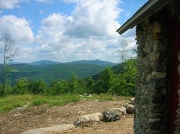 A scenic view from a stone building, showing rolling green hills under a partly cloudy sky. The foreground includes a rocky path and patches of grass, while the distant mountains create a tranquil backdrop. Green Mountain Trails mountain bike trail.