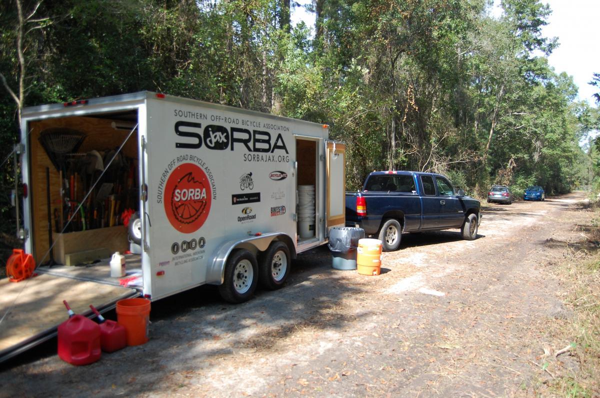 A blue pickup truck parked next to a trailer associated with the Southern Off-Road Bicycle Association (SORBA). The trailer is partially open, revealing various tools and equipment inside, with several containers and gas cans placed on the ground nearby. The setting is a wooded area along a dirt road. Tillie Fowler Regional Park mountain bike trail.