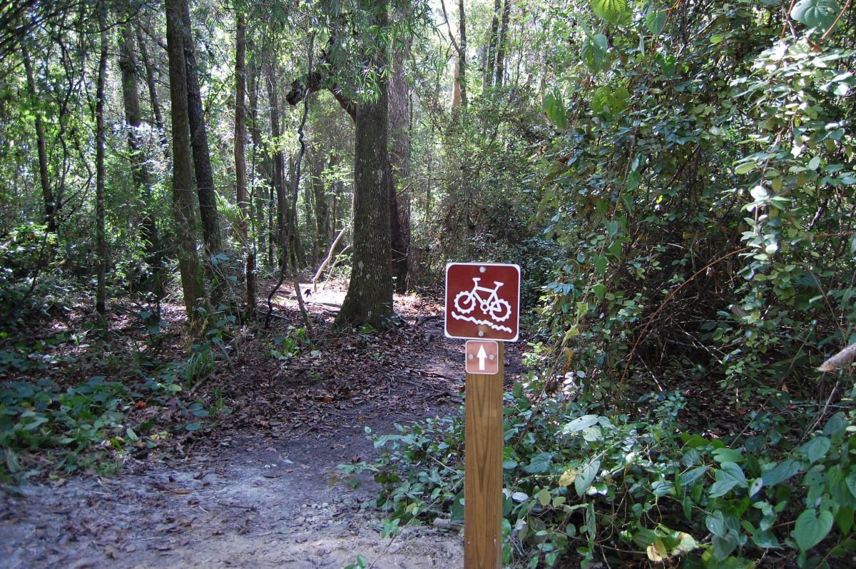 Sign indicating a bike trail entrance in a wooded area, with lush green foliage and tall trees surrounding a muddy path. The sign features a bicycle icon with an arrow pointing forward. Tillie Fowler Regional Park mountain bike trail.