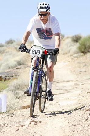 Specialized Crosstrail: A cyclist wearing sunglasses, a white t-shirt, and shorts rides a mountain bike on a dirt trail in a natural landscape. The cyclist has a race number (903) displayed on the front of the bike and is smiling as he navigates the terrain. Surrounding vegetation includes sparse shrubs and dry ground under a blue sky.