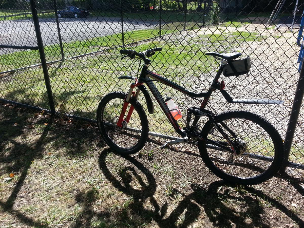 Trek Fuel EX 8: A black and red mountain bike leaning against a chain-link fence in a park, with a water bottle attached and shadows cast on the grass. A gravel path and playground equipment are visible in the background.