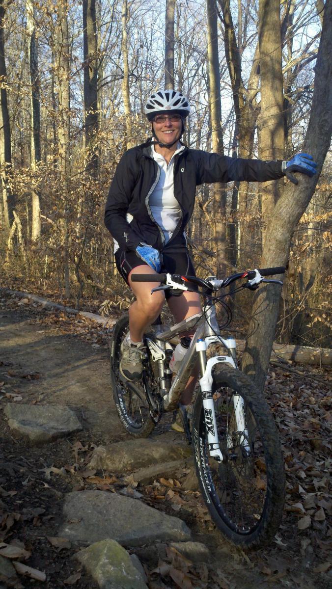 Trek Fuel EX 8: A person wearing a helmet and cycling gear smiles while sitting on a mountain bike on a rocky trail in a wooded area. The surrounding trees are bare, indicating it may be early spring or late fall. The cyclist has one hand resting on a nearby tree, and the ground is covered with fallen leaves and rocks.