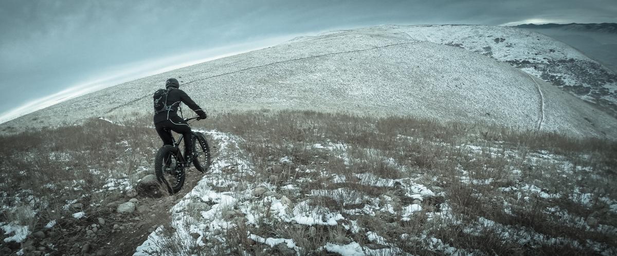 A cyclist riding a fat bike on a snowy mountain trail, surrounded by grassy terrain, under a cloudy sky. The cyclist is dressed in dark, fitted clothing and is navigating a rugged landscape. Green Mountain mountain bike trail.