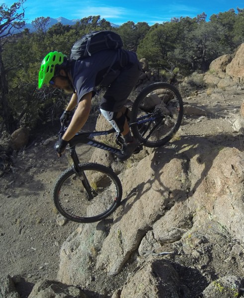 A mountain biker wearing a bright green helmet and a backpack rides over rocky terrain, navigating a steep incline in a wooded area.