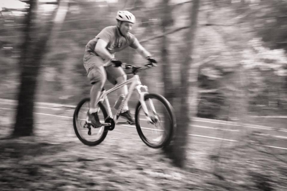 A person riding a mountain bike jumps through the air in a wooded area, surrounded by trees. The image is in black and white, capturing the motion and energy of the cyclist as they navigate the terrain. Governor's Creek mountain bike trail.