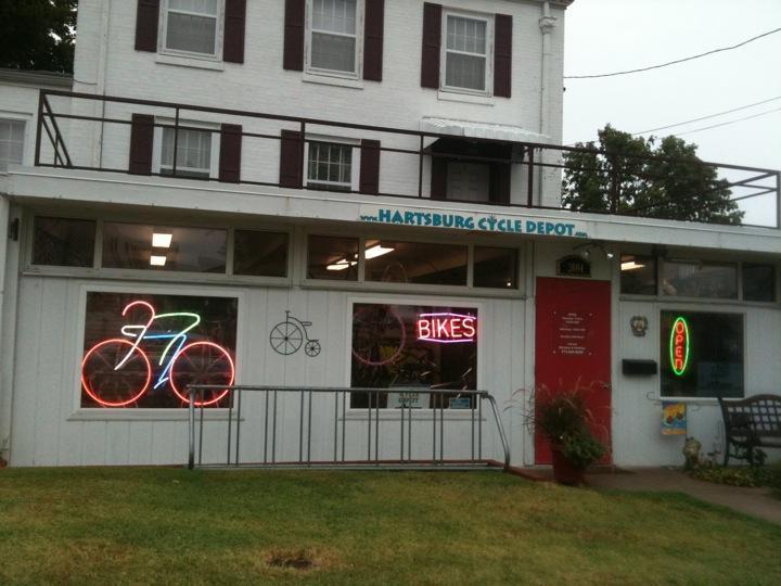The exterior of Hartsburg Cycle Depot, featuring a white building with multiple windows. Neon signs displaying a bicycle and "Bikes" illuminate the storefront. The sign above the entrance reads "Hartsburg Cycle Depot," and there is an "Open" neon sign visible. A grassy area is in front, with a bike rack and a small plant near the entrance.