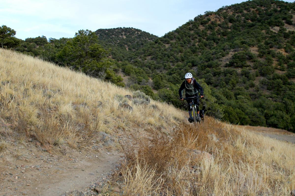 A person riding a mountain bike along a narrow dirt trail on a hillside, surrounded by dry grass and rocky terrain. Lush green hills and trees are visible in the background under a partly cloudy sky. North Backbone mountain bike trail.